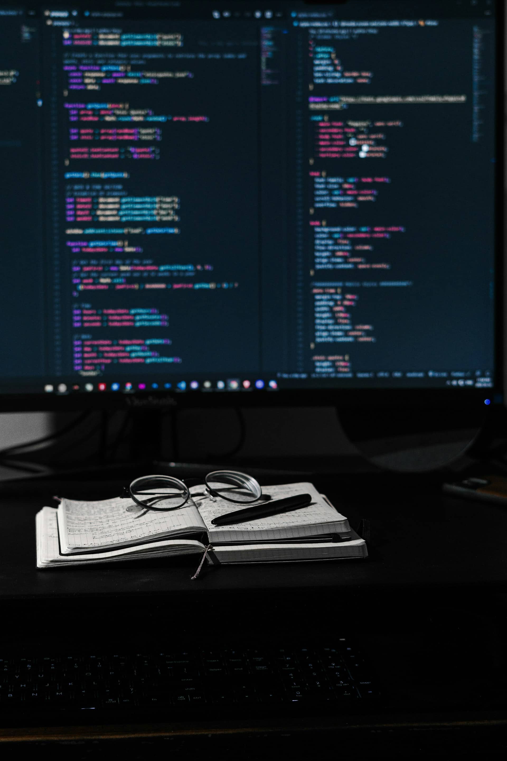 Компьютерный стол на котором лежит открытая книга, на книге лежат очки, а на заднем фоне монитор с html и css кодом.